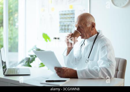 medico maschile in uniforme bevendo caffè e leggendo documenti medici su appunti seduta in ufficio ospedaliero. copia spazio Foto Stock