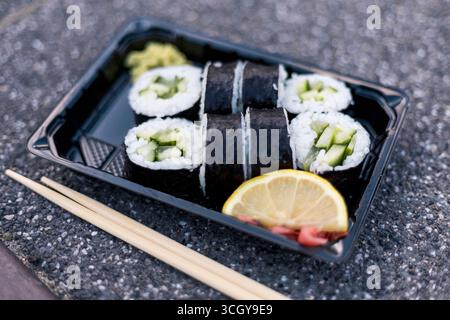 Deliziosi sushi roll ripieni di cetriolo fresco sono disposti in modo ordinato in un contenitore da asporto nero. Una fetta di limone e wasabi accompagnano il piatto Foto Stock