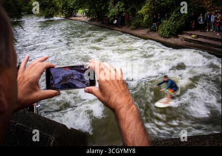 Monaco, Germania. 30 agosto 2025. Un uomo filma un surfista che cavalca l'onda artificiale di Eisbach nel giardino inglese. L'onda di Eisbach è un vero e proprio punto di riferimento per surfisti e spettatori in qualsiasi periodo dell'anno. Crediti: Peter Kneffel/dpa/Alamy Live News Foto Stock