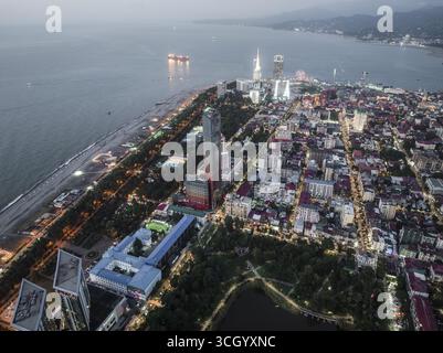 Vista aerea del paesaggio urbano illuminato che incontra la tranquilla costa del Mar Nero, con la Torre alfabetica e la ruota panoramica, Batumi, Adjara, Georgia. Foto Stock