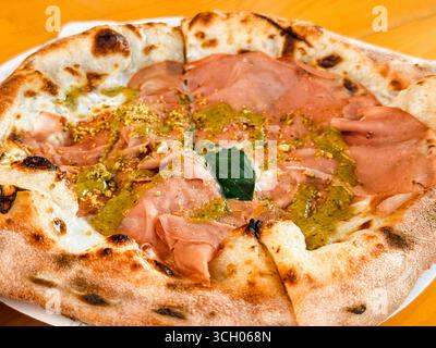 Pizza appena sfornata con mortadella, pesto, pistacchio e basilico su un tavolo di legno. Cultura gastronomica, gastronomia e tradizione culinaria Foto Stock