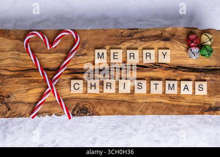 Saluto natalizio organizzato con piastrelle in legno che scrivono "buon Natale", accompagnato da canne di caramelle a forma di cuore e colorato jingle b Foto Stock