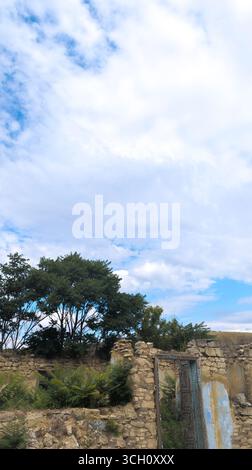 La natura recupera l'edificio abbandonato con pareti in pietra sbriciolata e portone in metallo arrugginito sotto il cielo blu nuvoloso Foto Stock