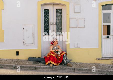 Castro Marim, Portogallo, 29 agosto 2025. Una donna vestita con un vivace abbigliamento da festival si prende una pausa mentre si siede di fronte a un edificio storico di CAS Foto Stock