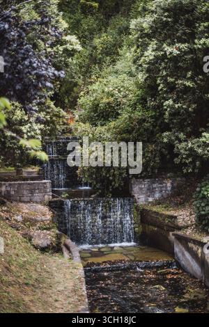 Piccola cascata circondata da lussureggianti alberi verdi e muri di pietra, con acqua limpida che scorre dolcemente attraverso un tranquillo parco, creando un Foto Stock