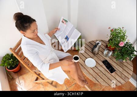 Una donna gode di un momento di pace su un balcone soleggiato, sorseggiando un caffè mentre legge un giornale. Circondata da vegetazione e fiori, si rilassa in un bagno Foto Stock