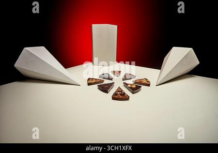 Una disposizione simmetrica con fette di cioccolato disposte in forma circolare su una superficie bianca, accentuate da forme a prisma angolari e bef impostato Foto Stock