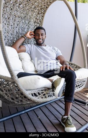 Seduto su una sedia sospesa, un uomo afroamericano sorridente con fiducia sul patio Foto Stock