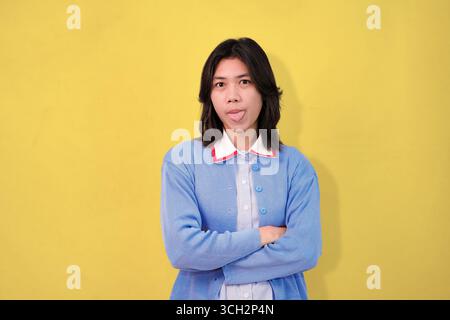 giovane che posa la lingua con un maglione blu su uno sfondo giallo brillante Foto Stock
