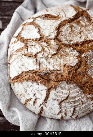 Dorato e croccante, il pane rotondo poggia su lino, la sua superficie crepata piena di carattere. Tradizioni fatte in casa e comfort di campagna. Vista dall'alto. Foto Stock