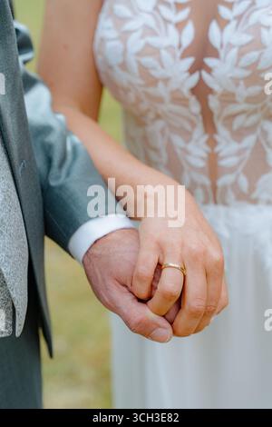 La sposa in un elegante abito in pizzo tiene le mani con lo sposo in un abito, mostrando una fascia nuziale dorata. L'immagine cattura un momento di connessione e unità Foto Stock