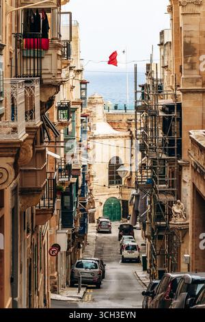 La Valletta, Malta - 10 aprile 2025: Strada stretta con balconi colorati e bandiera maltese nella capitale storica Foto Stock