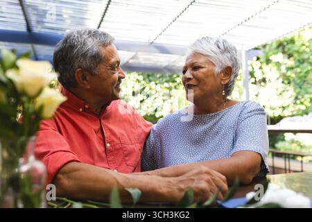Sorridente coppia anziana e diversificata che siede insieme al bar in giardino, godendo di una conversazione Foto Stock