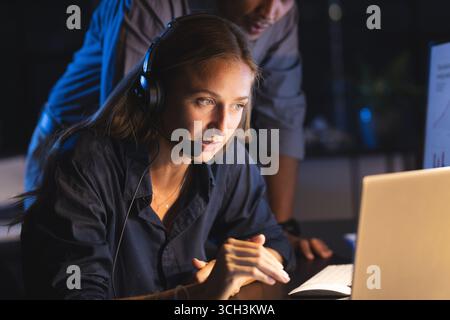 Donna d'affari concentrata che indossa un auricolare che lavora fino a tardi con un laptop in ufficio Foto Stock
