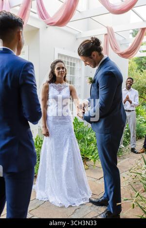 Sposa e sposo si scambiano anelli all'aperto, circondati da amici gioiosi Foto Stock