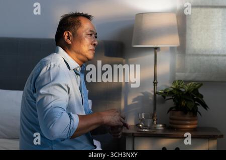 Uomo anziano seduto sul letto di casa, riflettendo nella luce soffusa della sera Foto Stock