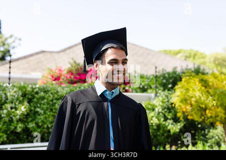 Uomo indiano sorridente mentre è in piedi davanti al cortile con berretto e abito nero in mortarboard Foto Stock