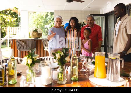 Riunirsi intorno a un tavolo, una famiglia multigenerazionale che sorride durante un pasto in giardino insieme Foto Stock