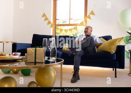 Uomo anziano che si rilassa sul divano a casa durante le festività natalizie con palloncini Foto Stock