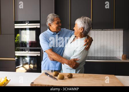 Una coppia senior che abbraccia la cucina moderna, sorride, si gode la compagnia a casa Foto Stock