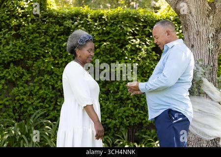 Coppia senior che si scambiano anelli in giardino, celebrando l'amore e l'impegno all'aperto Foto Stock