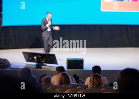 Relatore che presenta alla conferenza aziendale con il pubblico seduto e sfondo digitale dello schermo. Ambiente professionale. Foto Stock