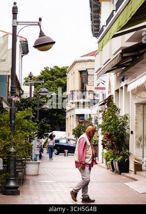 Limassol, Cipro, 04-23-2025. Un vecchio che cammina in una vivace strada pedonale caratterizzata da edifici in vecchio stile e verde Foto Stock