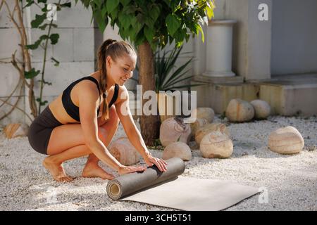 La donna si prepara allo yoga all'aperto con un tappetino in un cortile con giardino soleggiato, spazio pulito per il fitness benessere e concetti di consapevolezza Foto Stock