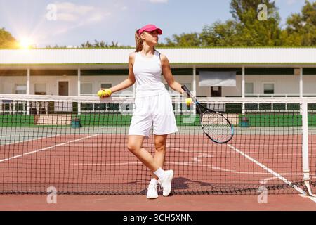 Giovane tennista donna sicura di sé con un vestito bianco e una visiera rosa appoggiata su rete con racchetta e palle da tennis. Foto Stock