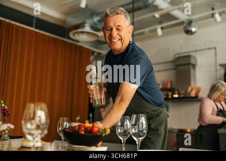 Vista ad angolo basso dell'anziano sorridente che tiene in mano una bottiglia d'acqua mentre prepara il tavolo durante la lezione di cucina Foto Stock