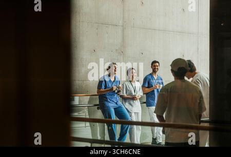 Uomini e donne felici professionisti medici che si godono una pausa caffè in corridoio visto attraverso la parete di vetro Foto Stock
