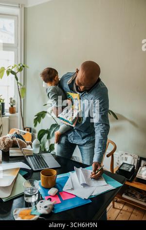 Libero professionista che porta un bambino che parla con uno smartphone mentre scrive appunti su carta a casa Foto Stock