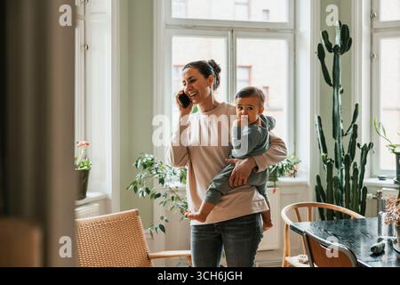 Donna felice che porta il figlio mentre parla con uno smartphone a casa Foto Stock