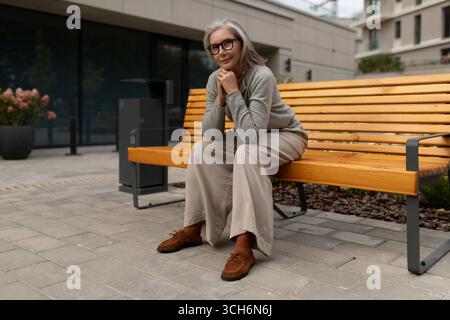 Donna anziana seduta comodamente su una panchina di legno in un ambiente moderno all'aperto durante il giorno Foto Stock