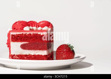 Una deliziosa fetta di torta di velluto rosso condita con cremosa glassa e accompagnata da una fragola fresca, presentata su un piatto bianco, perfetta per la panificazione Foto Stock