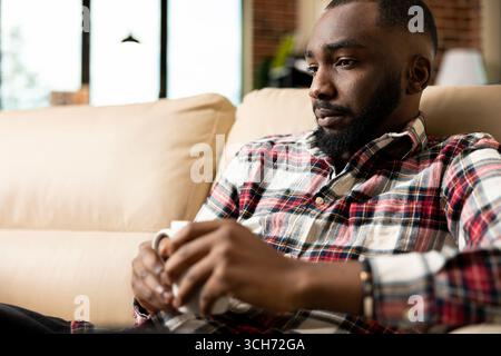 Il contabile remoto si siede sul divano con il caffè in mano, riflettendo profondamente sulle strategie per migliorare le finanze aziendali. Consulente aziendale che si prende una pausa dai progetti, bevendo una tazza di tè nel soggiorno. Foto Stock