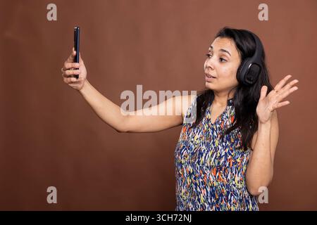Bella modella femminile che indossa le cuffie si aggancia al cellulare mentre sorride. Graziosa donna indiana con cuffie wireless, gode di una videochiamata con la famiglia, in piedi su uno sfondo isolato. Foto Stock