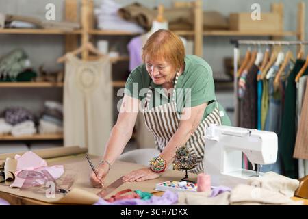 La donna sartoria lavora in un laboratorio di cucito e crea modelli di vestiti su carta. Personalizza con un metro a nastro per creare un design alla moda Foto Stock
