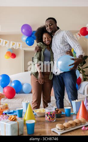 Coppia che abbraccia con palloncini e decorazioni per feste a casa per festeggiare il compleanno Foto Stock