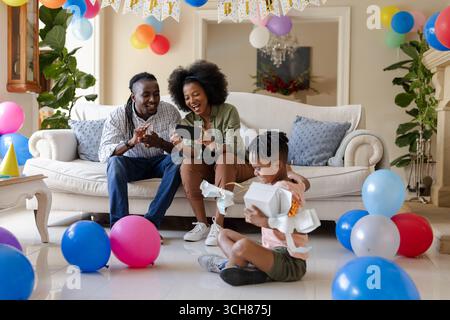 Alla festa, la famiglia festeggia il compleanno a casa, i genitori sorridono mentre il bambino apre il regalo Foto Stock