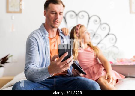Padre che usa lo smartphone mentre la figlia guarda lontano seduto sul letto di casa Foto Stock