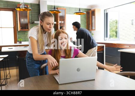 Le ragazze adolescenti guardano con entusiasmo lo schermo del notebook nella cucina moderna, celebrando il successo Foto Stock