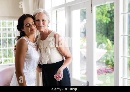 Abbracciando una donna anziana, la sposa sorride calorosamente in una stanza illuminata dal sole, festeggiando insieme Foto Stock