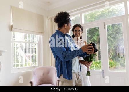 La sposa regge il bouquet sorridente allo sposo nel luminoso soggiorno, condividendo momenti di gioia Foto Stock
