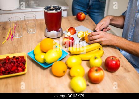 Primo piano di una coppia adulta che prepara il frullato in cucina. Stanno mescolando vari tipi di frutta nella centrifuga elettrica. L'uomo sta affettando l'arancia. Foto Stock