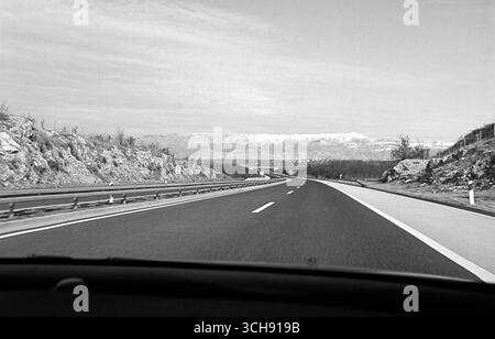 Foto POV in bianco e nero scattata dal sedile del passeggero di un'auto in movimento, che mostra un'autostrada aperta che si estende verso la lontana catena montuosa di Velebit. Foto Stock
