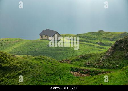 Casa di montagna isolata su colline verdi ondulate, circondata da natura tranquilla e luce soffusa. Paesaggio tranquillo che simboleggia la solitudine e la serenità Foto Stock