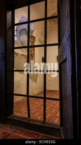 Vista di una vecchia cella della prigione con pareti in gesso sbriciolate e pavimento in piastrelle di terracotta visto attraverso barre di metallo. Foto Stock