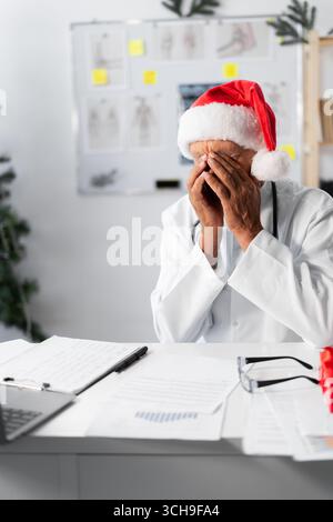 Anziano medico anziano nel cappello di babbo natale si sente stanco lavorando sul turno sembra esausto e coprirsi il viso con le mani alla vigilia di natale . Copia spazio Foto Stock