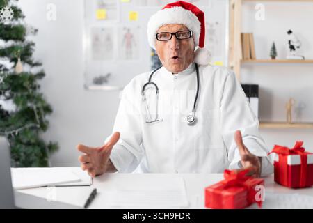 Anziani Dottore in cappello di Babbo Natale durante il turno della vigilia di natale con un'espressione facciale stupita e scioccata. copia spazio Foto Stock